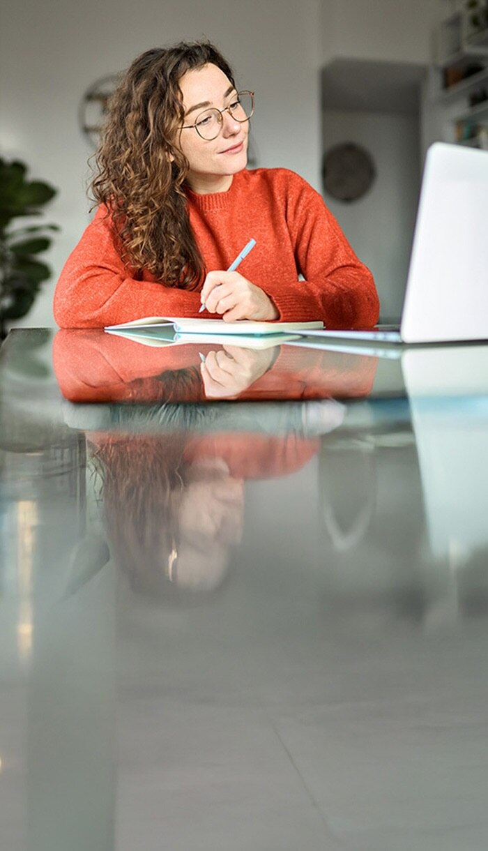 A woman in a red sweater curiously looks at her laptop screen and takes notes in her notebook about Norton Editorial Policies.